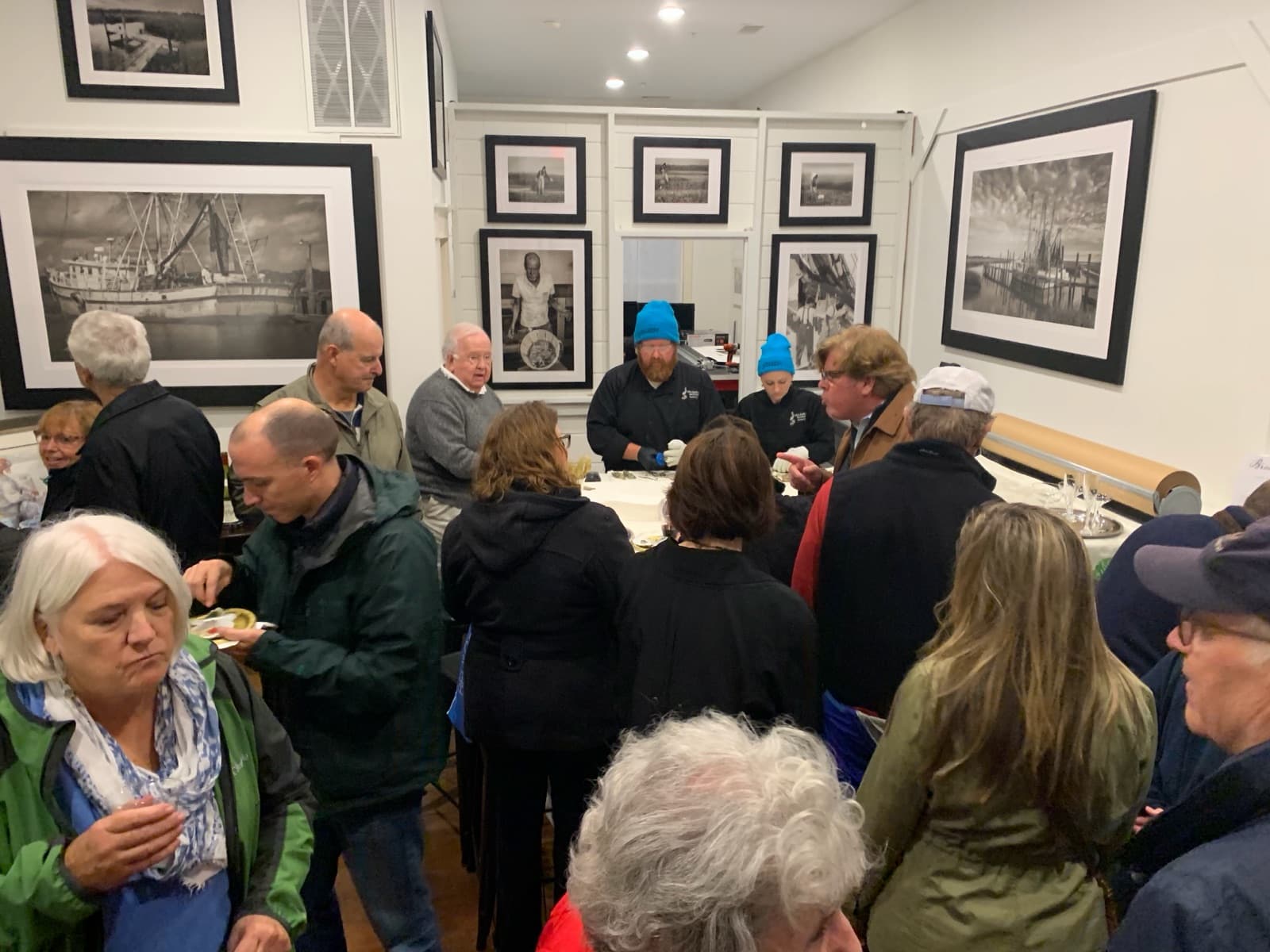 A crowd at the Branning Fine Art opening, surrounded by framed black and white photographs of shrimp boats and the Lowcountry
