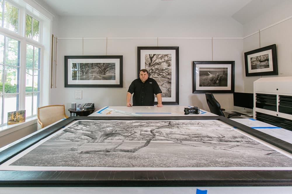 Andrew Branning in the Branning Fine Art gallery behind a large framed black and white print of a live oak